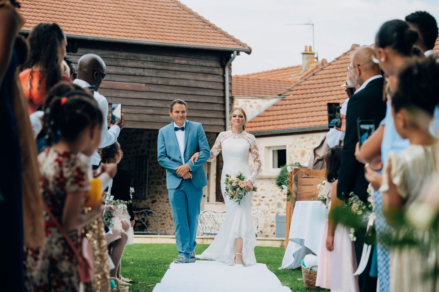 Bride in white gown walking down the aisle with escort at a rustic outdoor wedding ceremony.