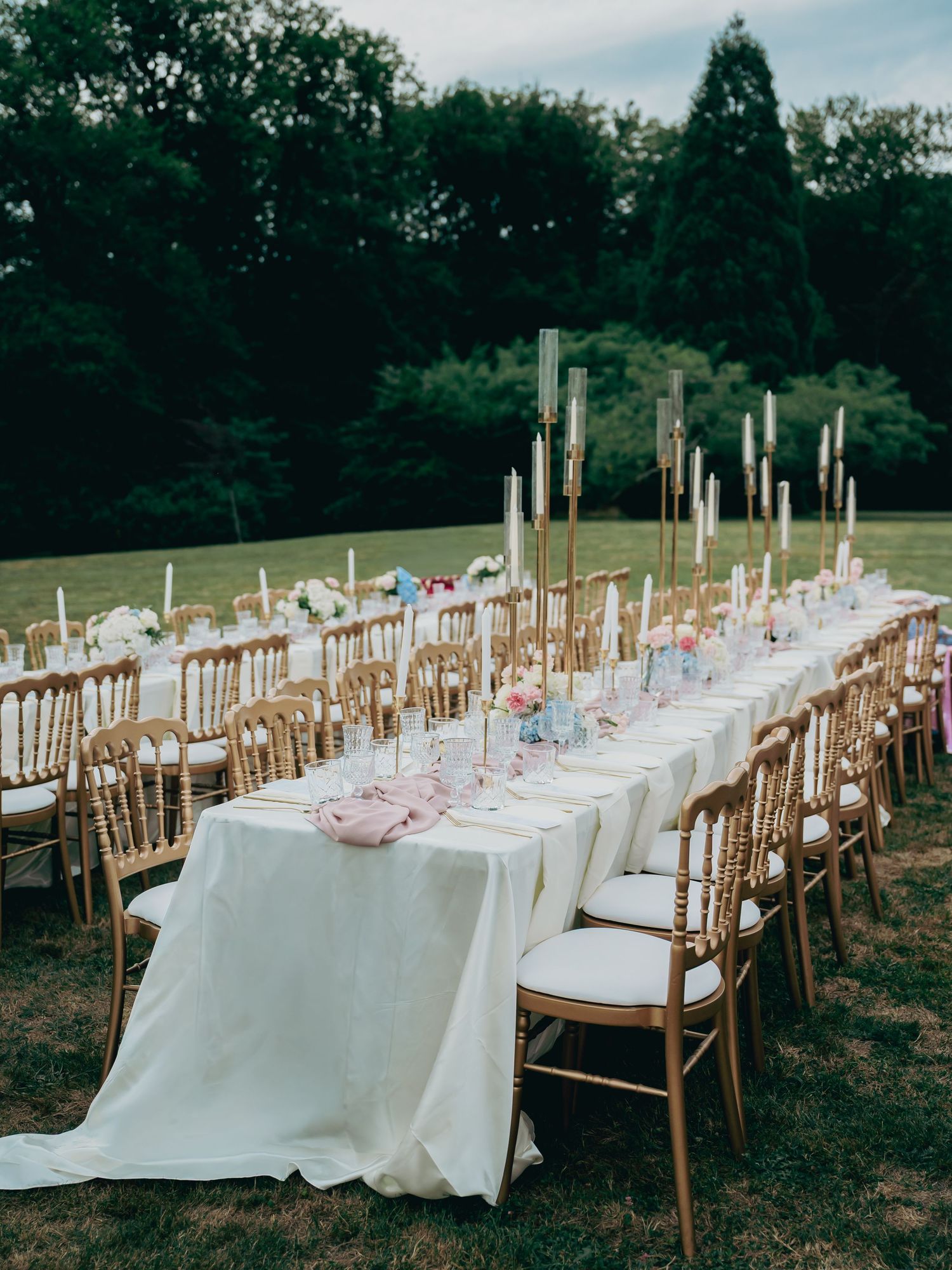 Stunning outdoor wedding reception with long white tables, gold chairs, and tall candles.