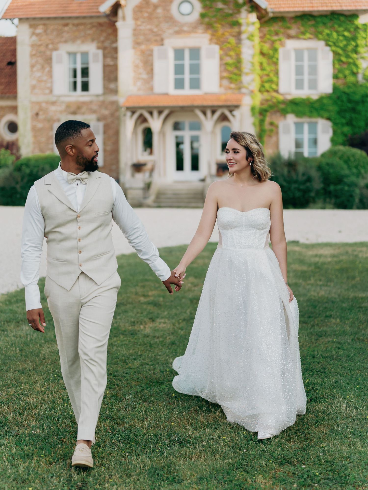 Interracial couple walks hand in hand on green lawn before a charming French country estate on their wedding day.