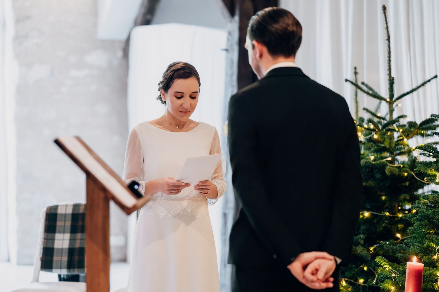 Bride reads wedding vows from paper at altar beside Christmas tree during winter ceremony.