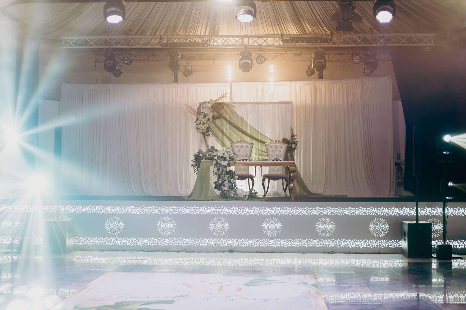 Grand wedding reception stage with ornate gold throne chairs and white draped backdrop with lights.