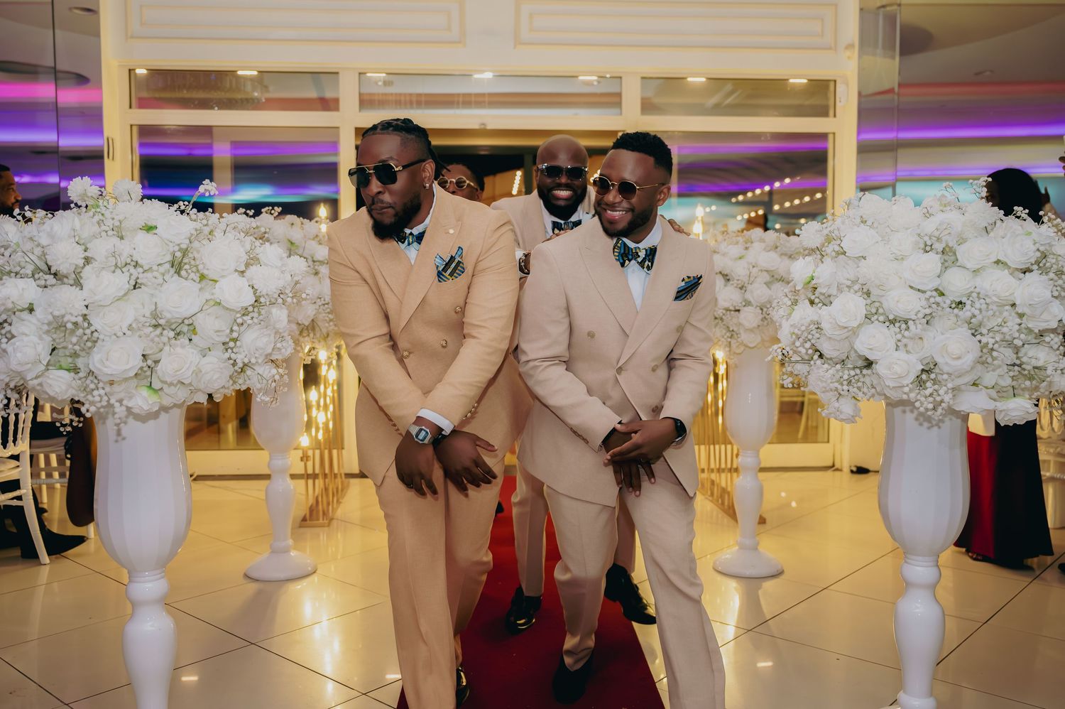 Two men in matching champagne suits with teal bow ties posing beside white floral arrangements at wedding reception.