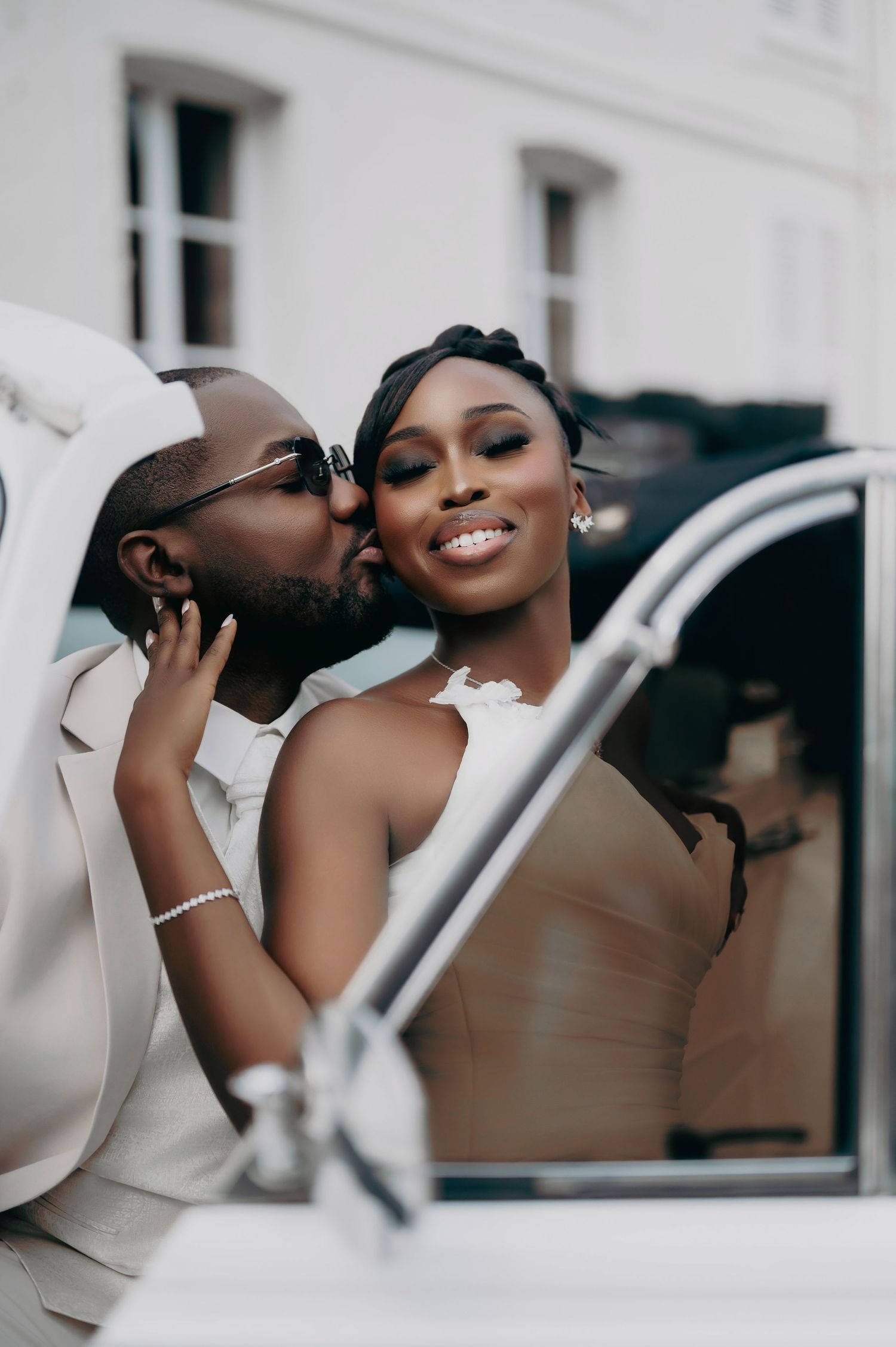 Groom kisses bride on cheek as she smiles near a white vintage car, reflected in the mirror.