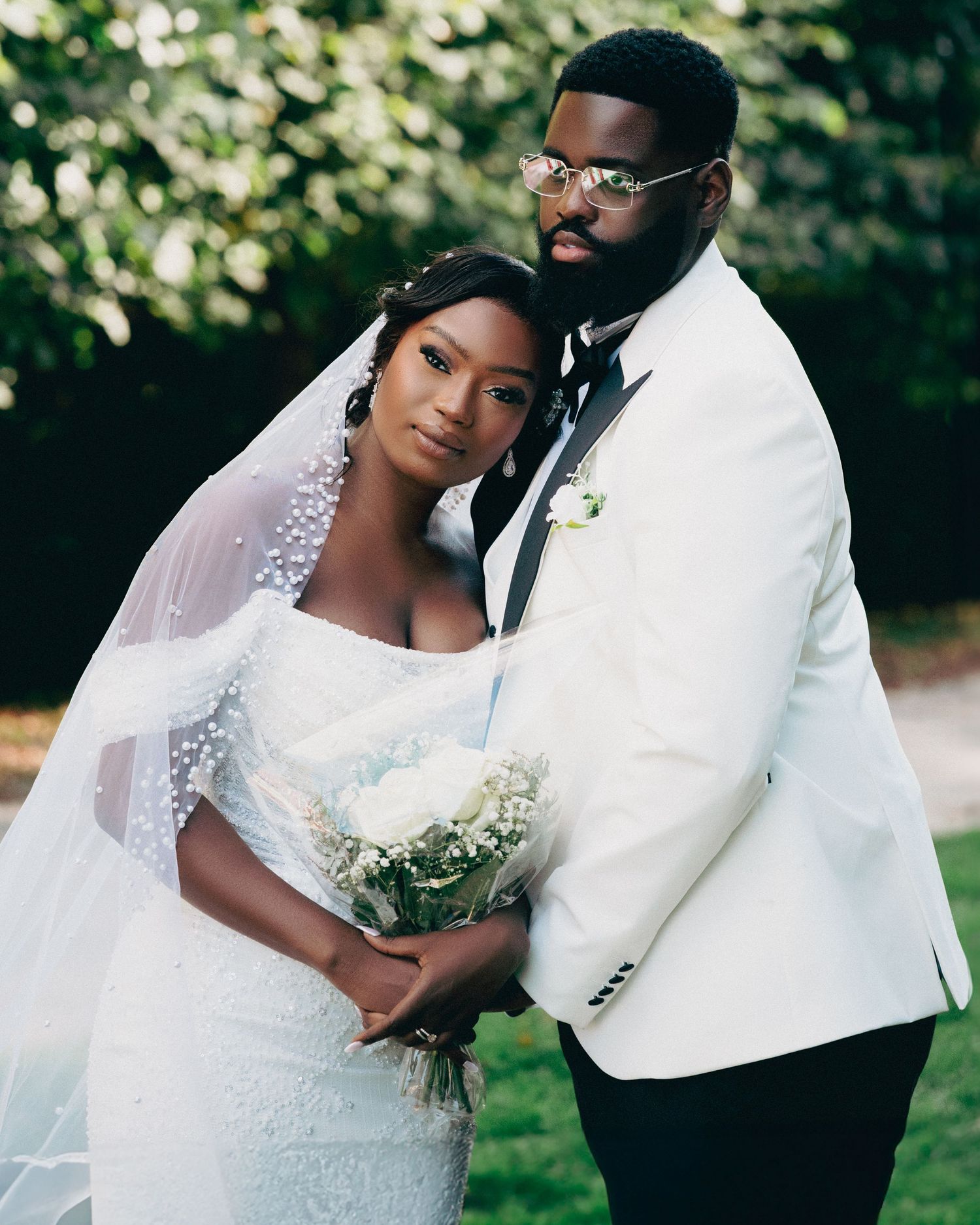 Black bride and groom posing outdoors, bride holding white bouquet, groom in white suit with glasses.
