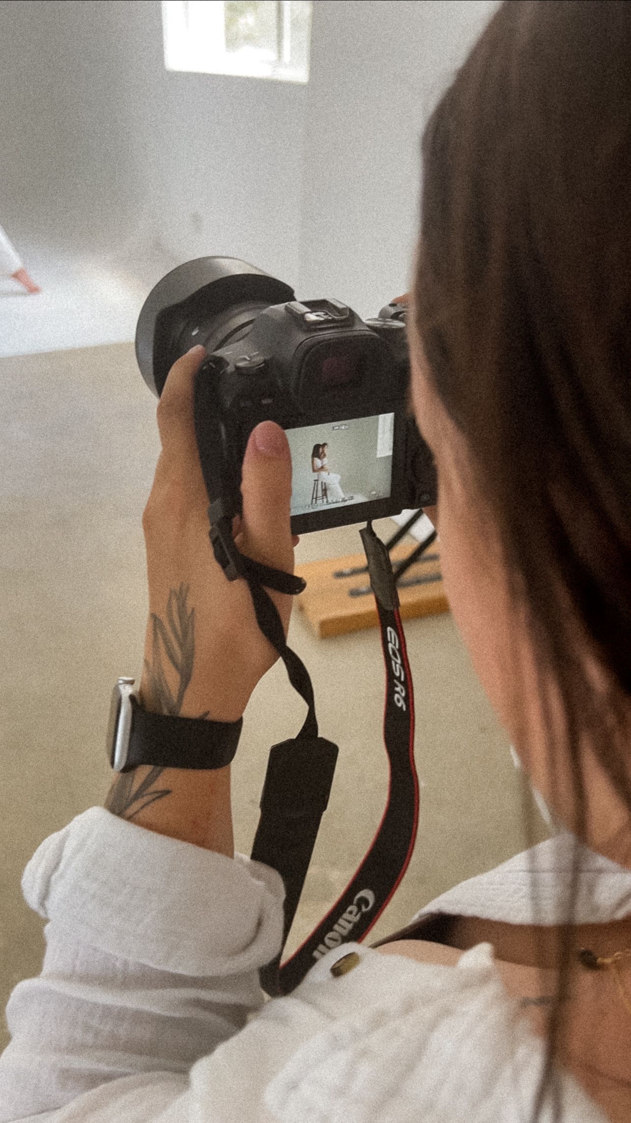 Photographer with visible arm tattoo holds Canon camera while reviewing images on LCD screen during indoor photo session.
