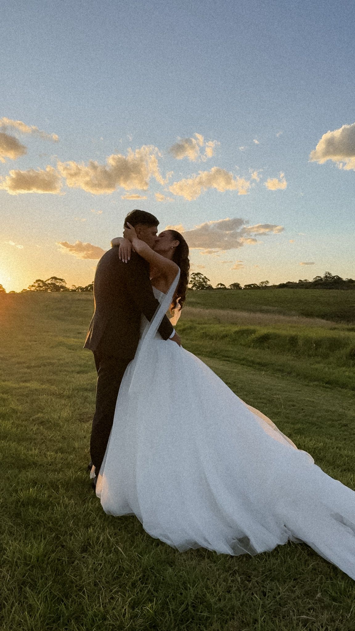 Newlyweds embrace in a grassy field during golden hour sunset.