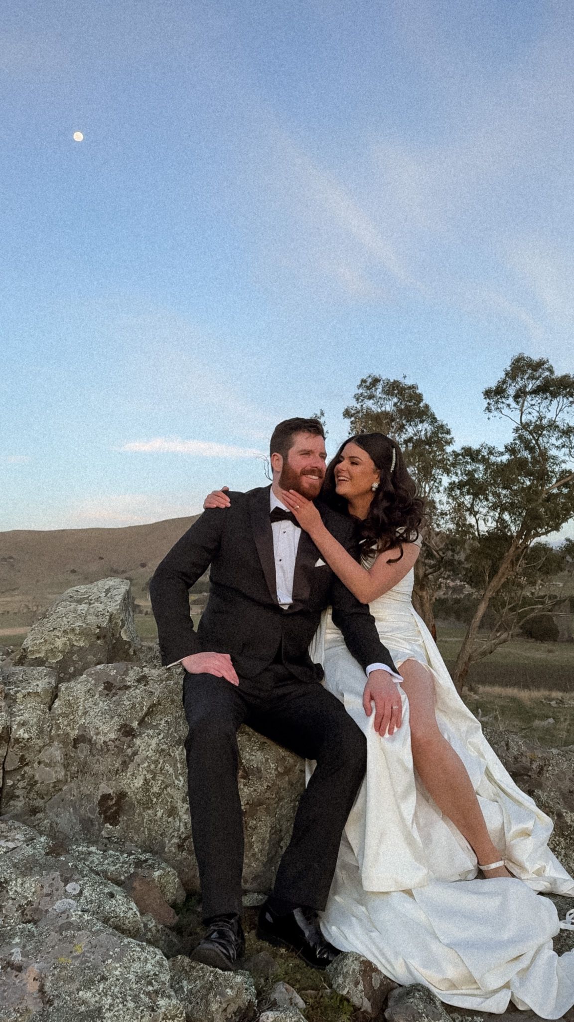 A couple in wedding attire poses playfully on rocky terrain at sunset.