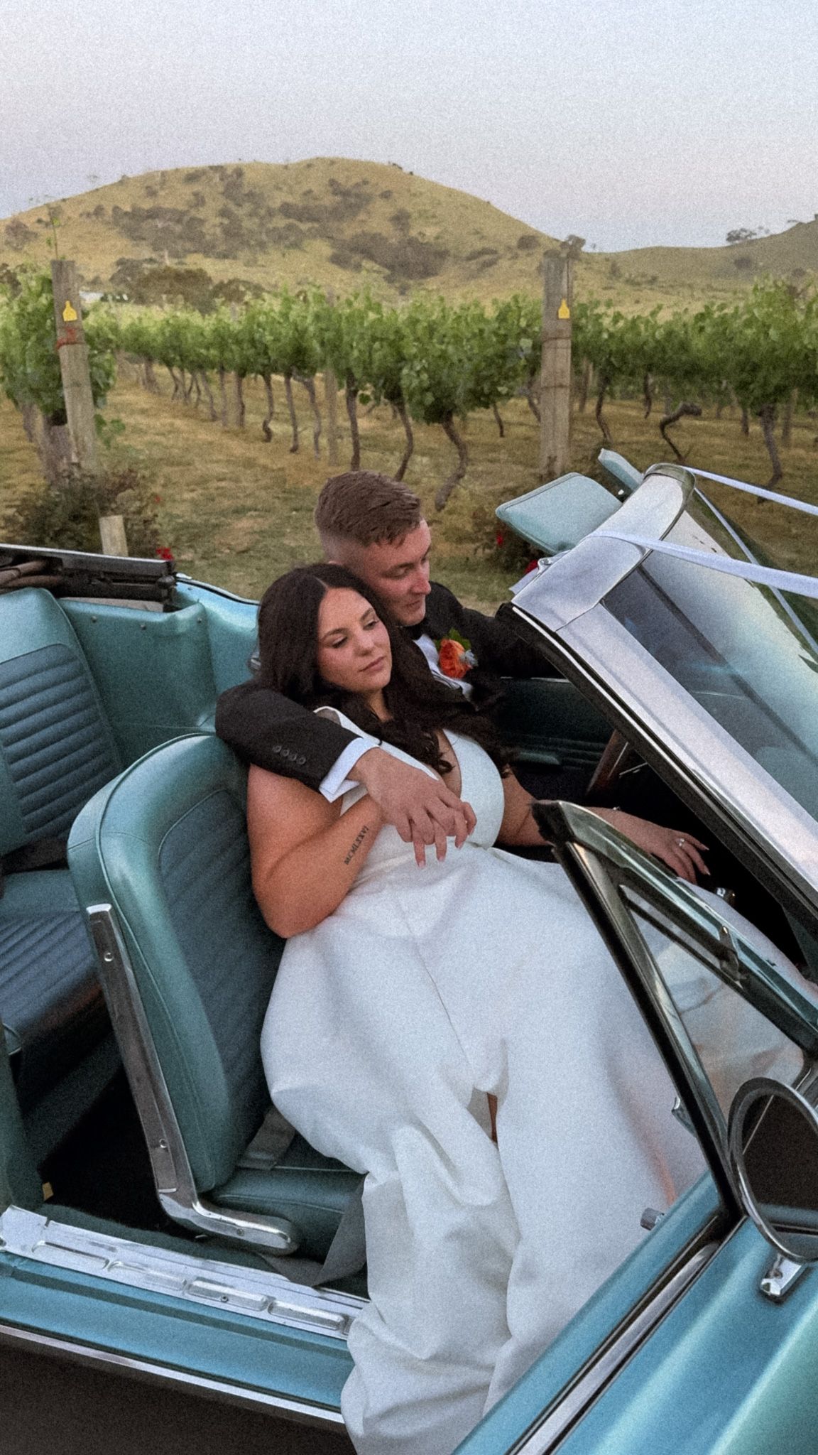 Newlyweds share an intimate moment in a vintage blue convertible near a vineyard.
