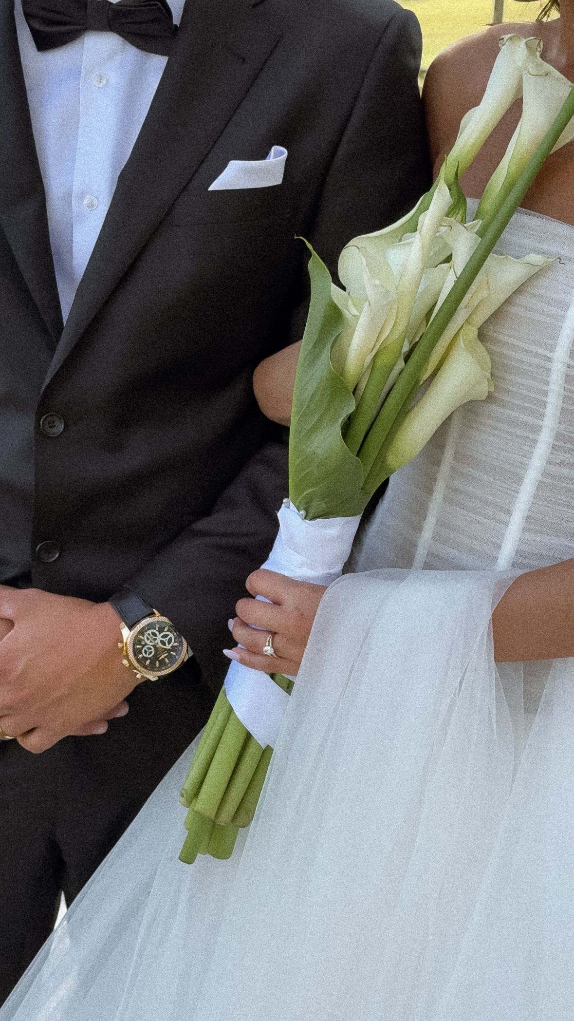 A wedding couple in formal attire with the bride holding a bouquet of white calla lilies against a white background.
