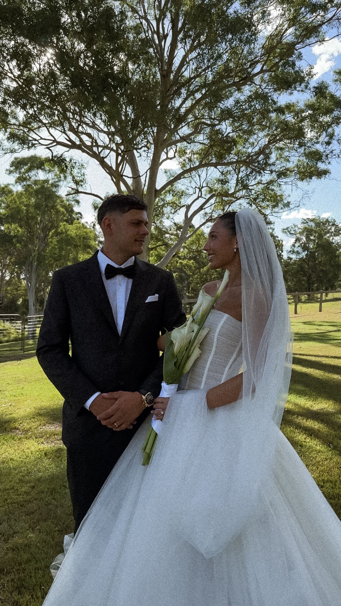 A bride in a white ball gown and veil stands with her groom in a black tuxedo under a large tree in a park setting.
