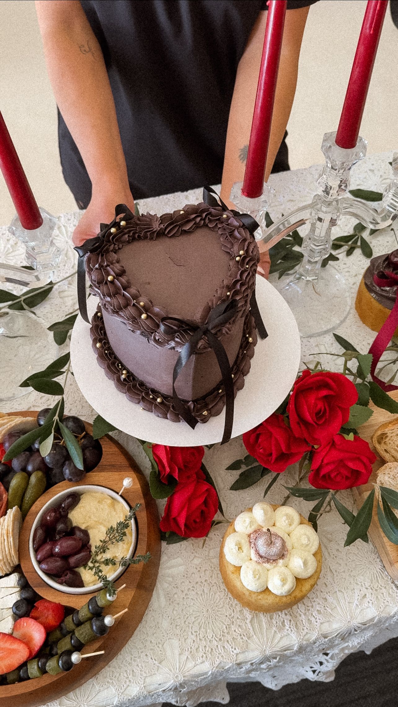 Heart-shaped chocolate cake with red roses and dessert platter on white lace tablecloth.