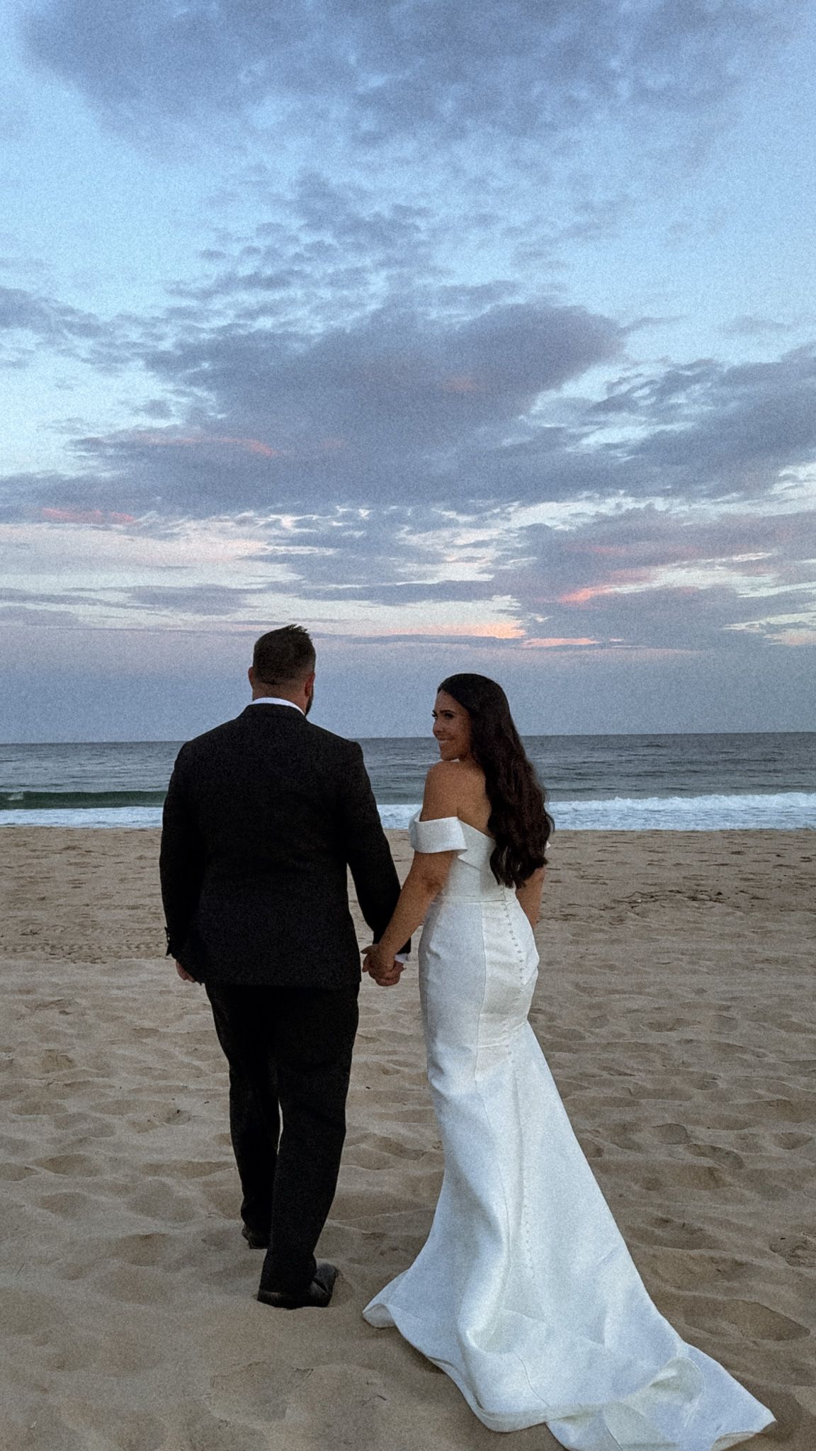Couple walking along beach at sunset with wedding attire.