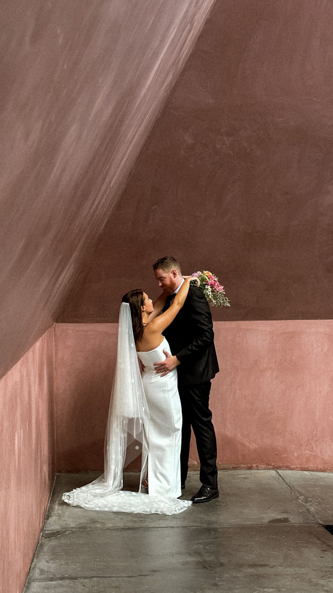 A couple shares an intimate moment against a pink concrete wall in wedding attire.