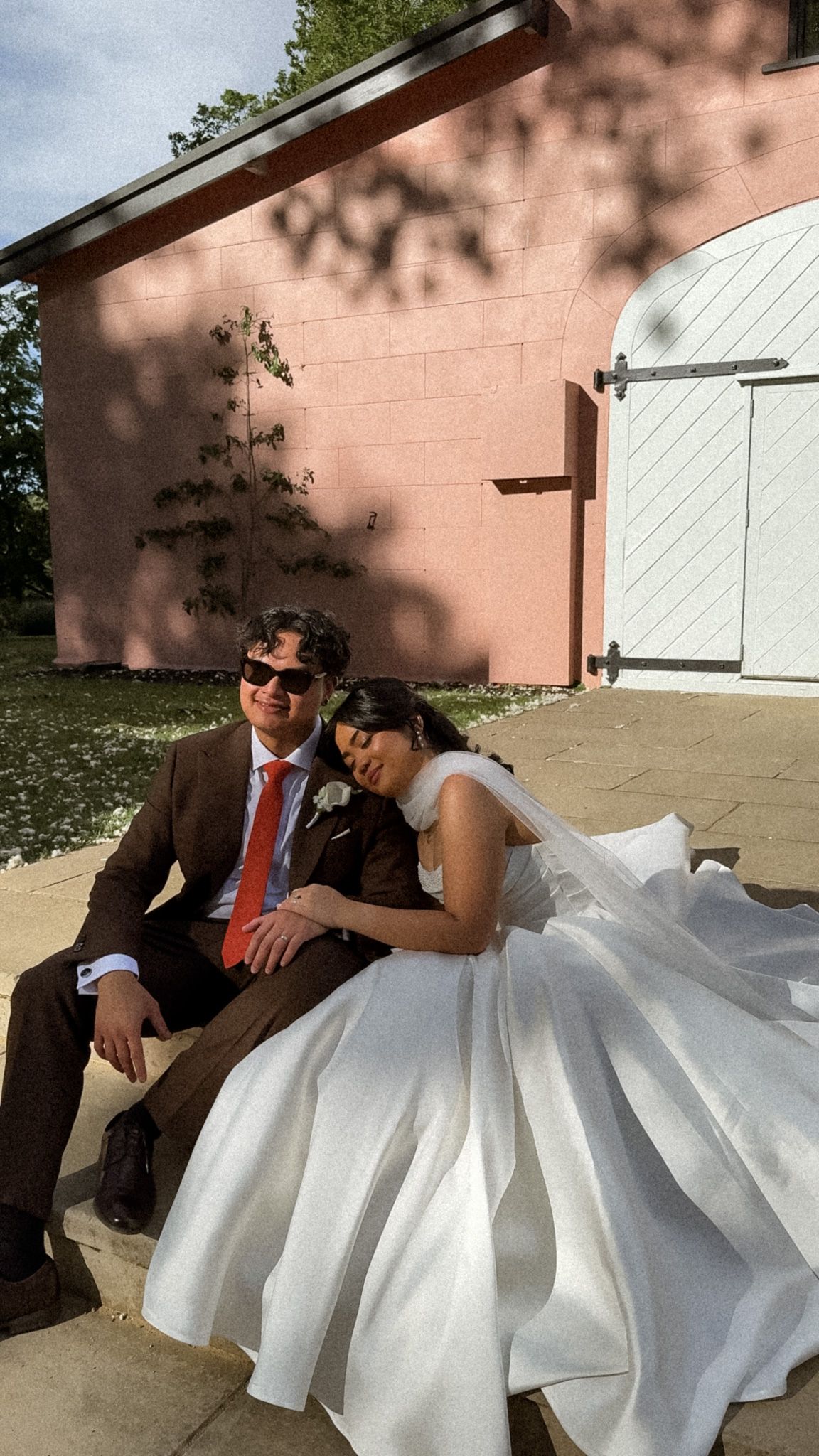 A couple in wedding attire resting on steps against a pink wall with white shutters.