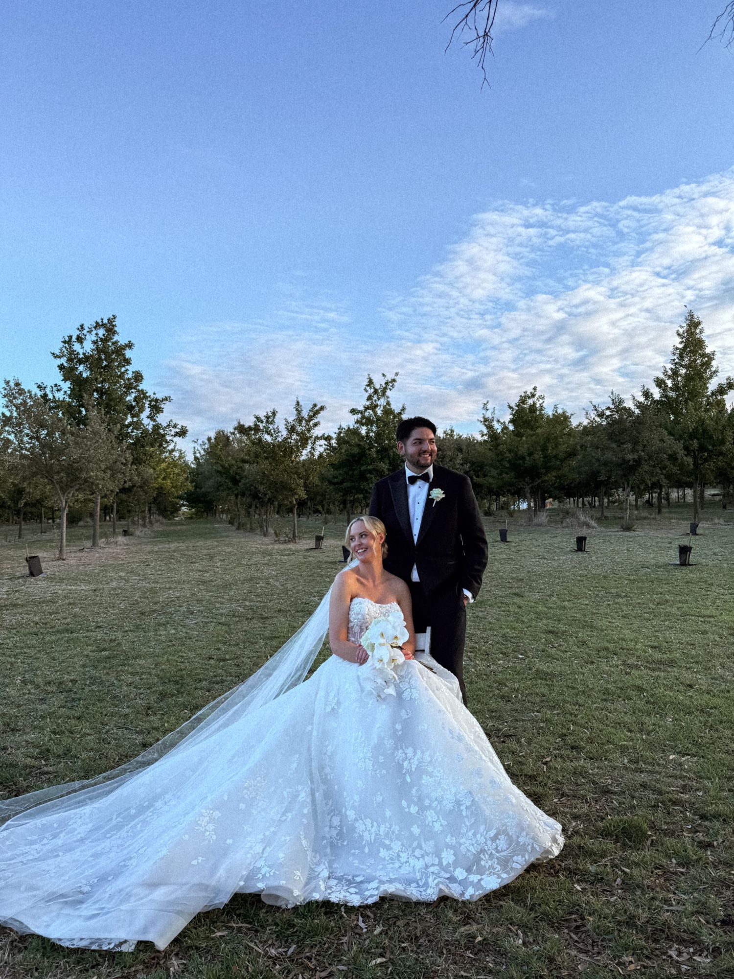 A couple in wedding attire poses together on a grassy field under a blue sky.
