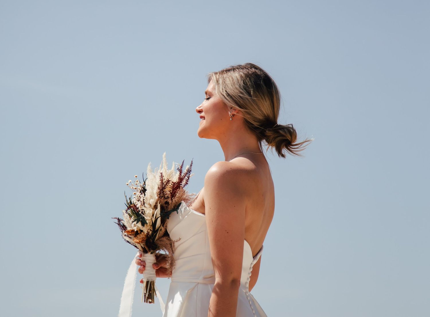Bride in lace dress stands against blue sky holding bouquet.