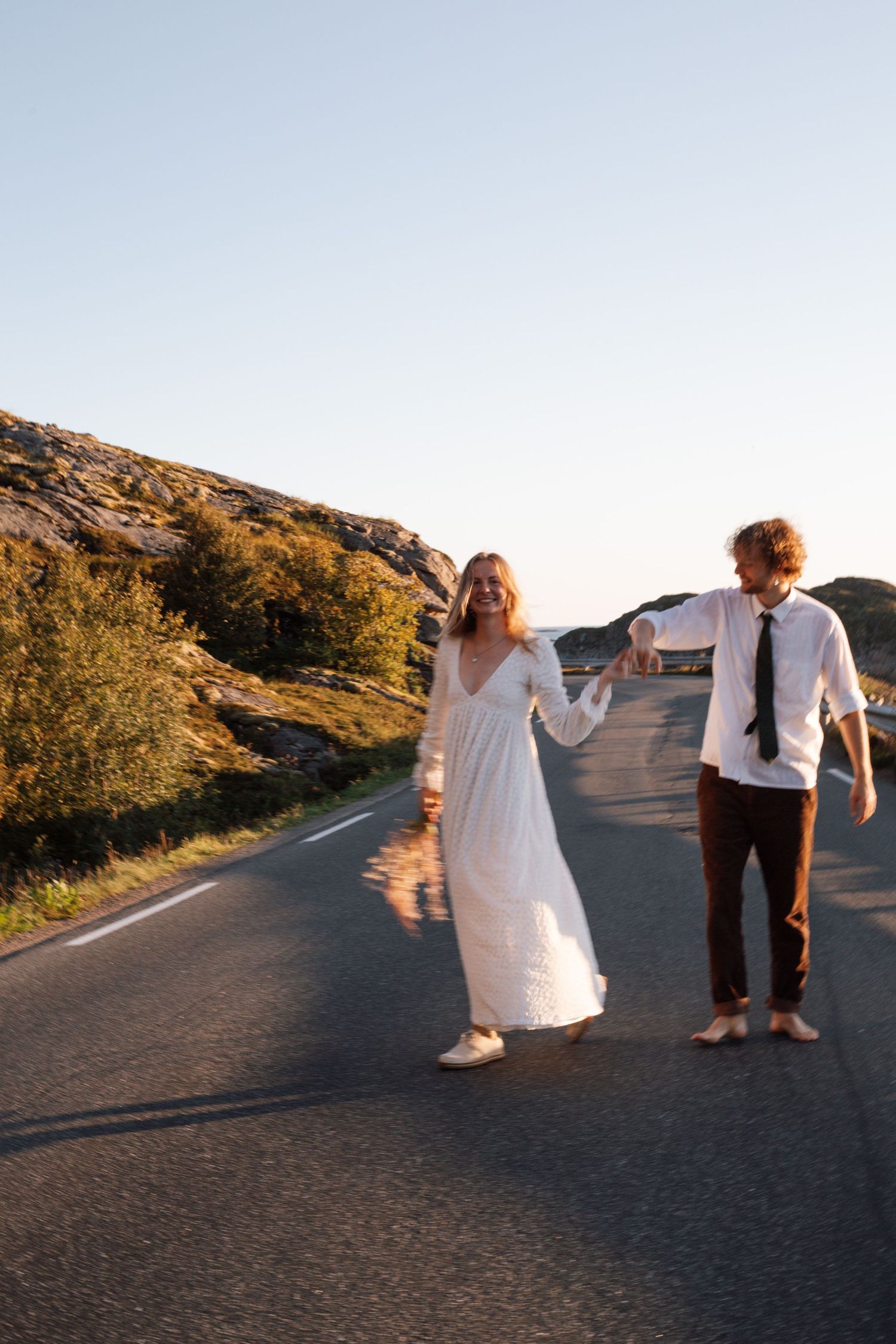 A couple in wedding attire walks playfully along a scenic mountain road at sunset.