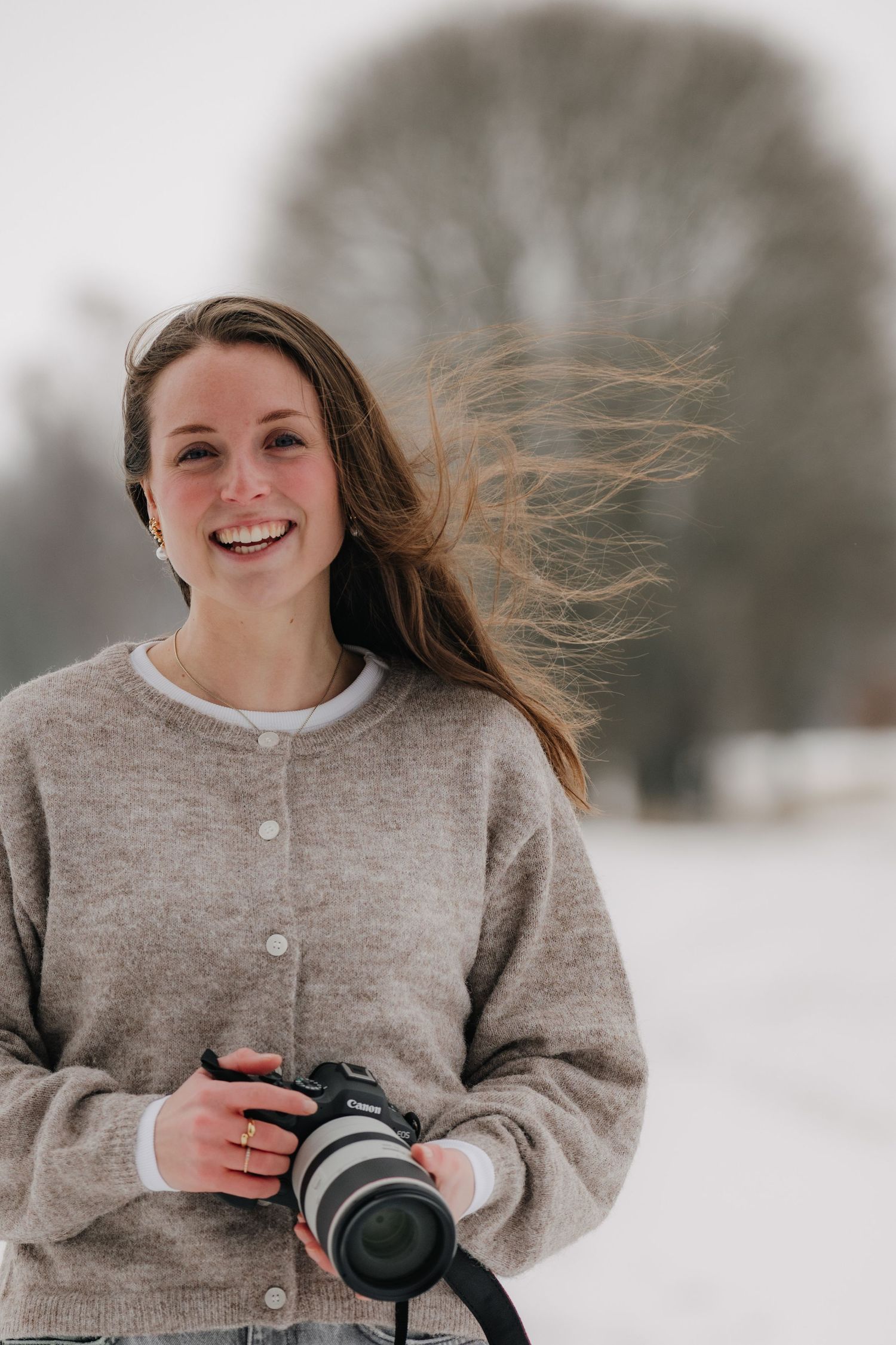 Photographer wearing grey cardigan smiles while holding camera in snowy setting.