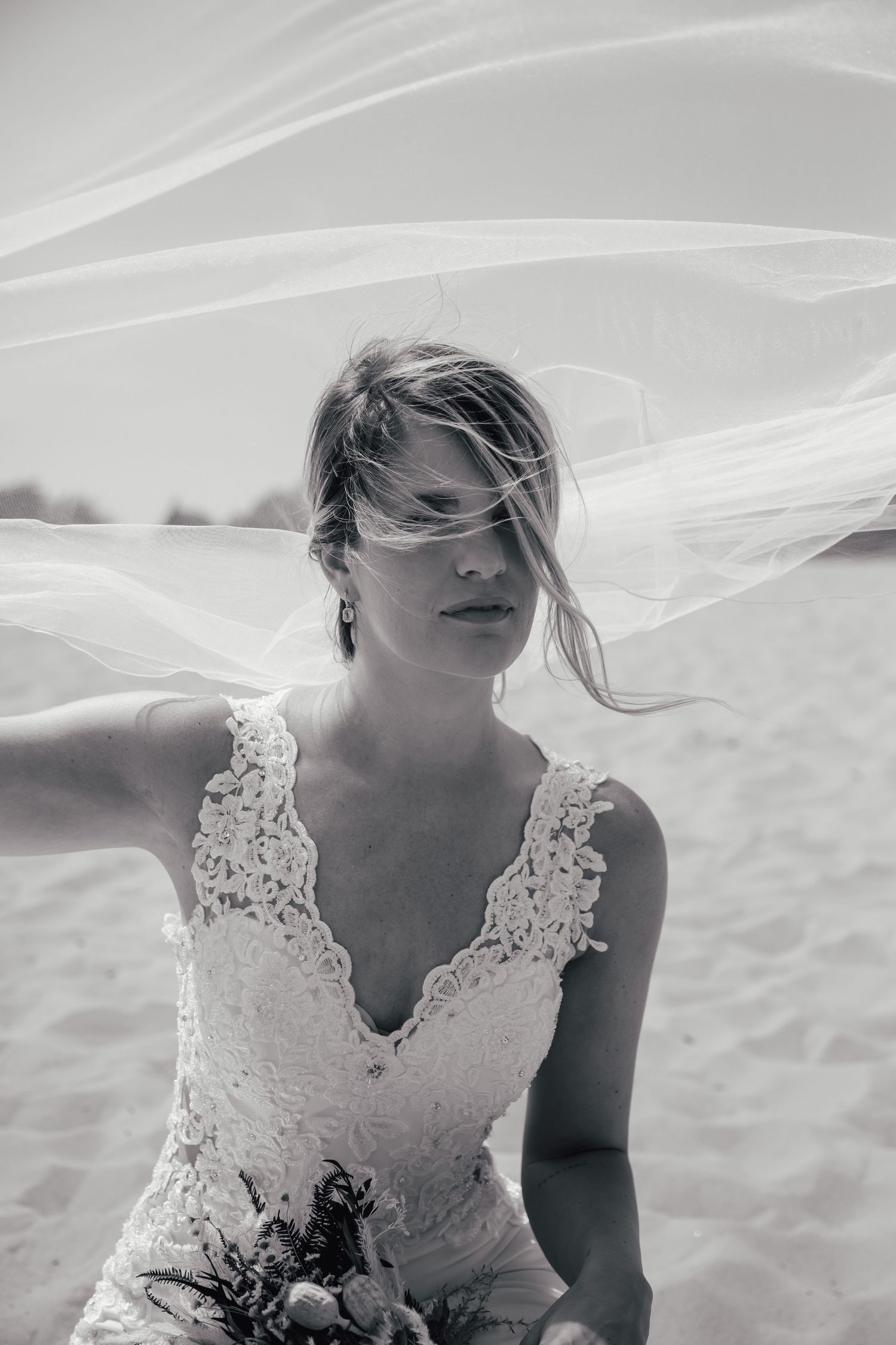 A bride in a lace wedding dress stands outdoors with a delicate veil flowing in the wind.