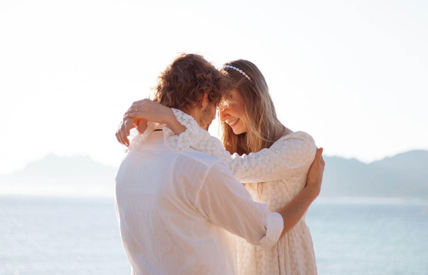 Intimate embrace between couple in white clothing against ocean backdrop.