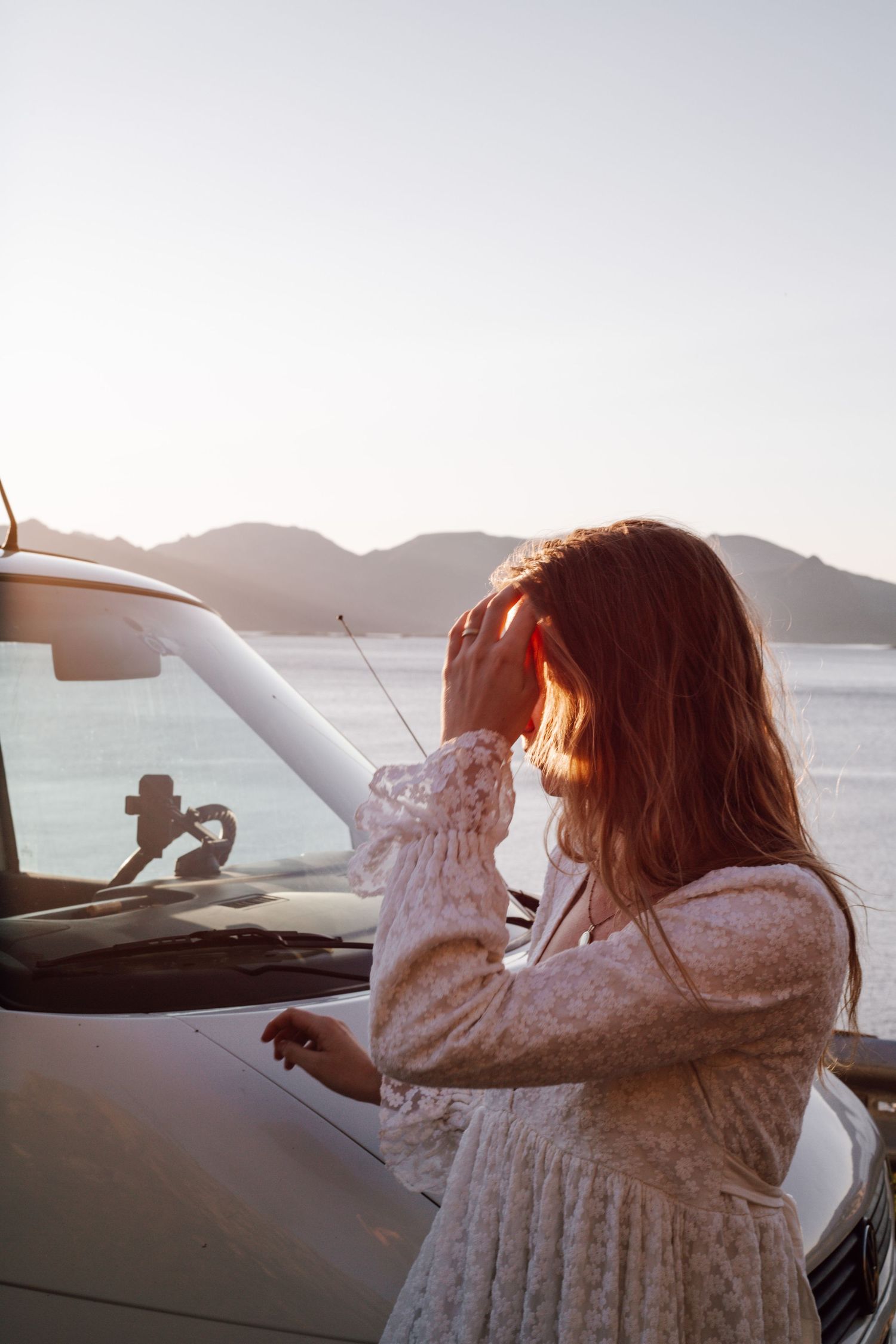 Woman in white dress poses playfully by car with mountain and ocean view.