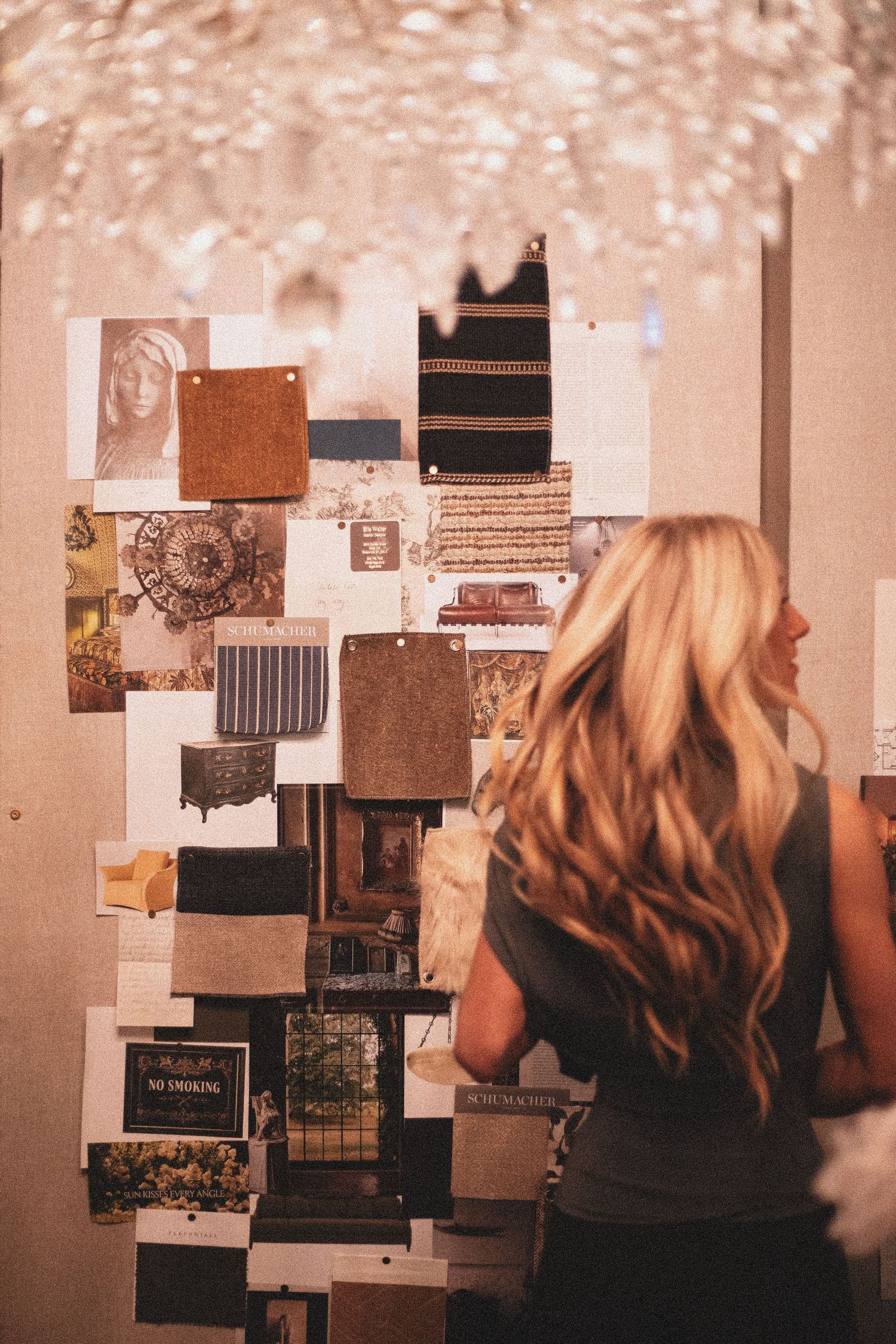 Blonde woman viewing an interior design mood board with fabric swatches and decor images under a crystal chandelier.