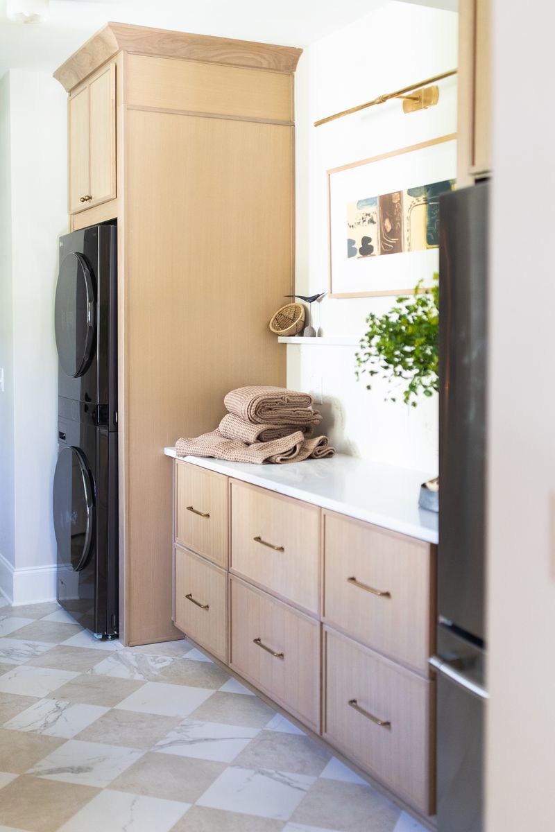 Modern laundry room with light wood cabinets, marble countertop, stacked washer-dryer, gold hardware, and checkered tile floor.
