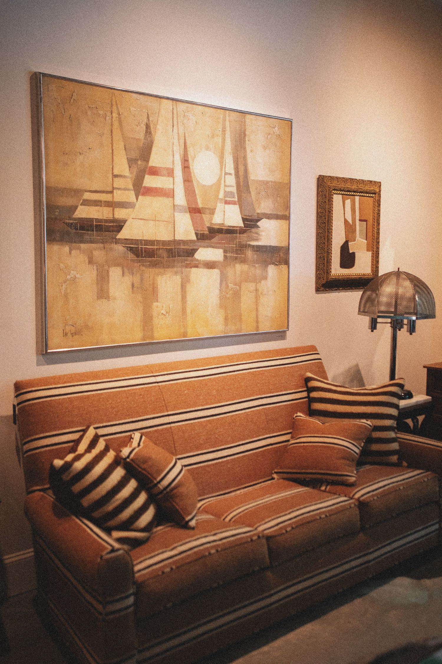 Brown striped sofa with decorative pillows beneath a sailboat painting and small framed artwork, beside a vintage lamp.