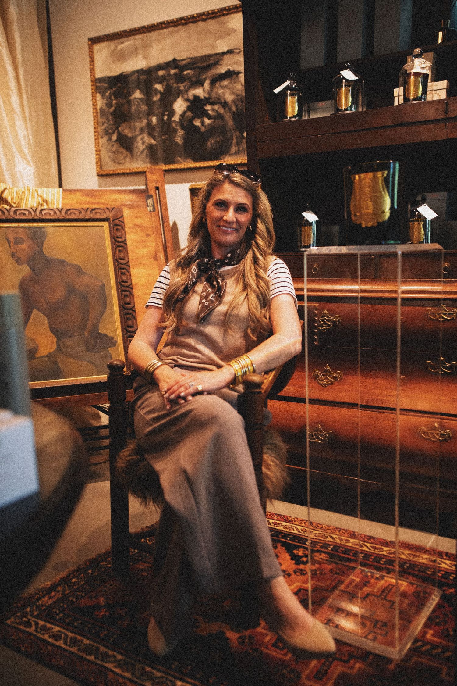 Woman smiling in chair surrounded by artwork, perfume bottles, and antique furniture in an elegant room.