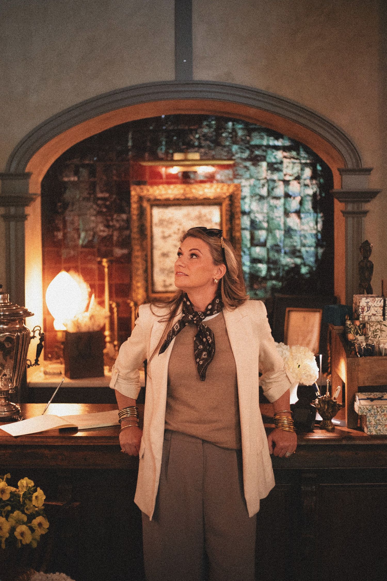 Elegant woman in white blazer and silk scarf stands in warmly lit vintage interior with arched fireplace.