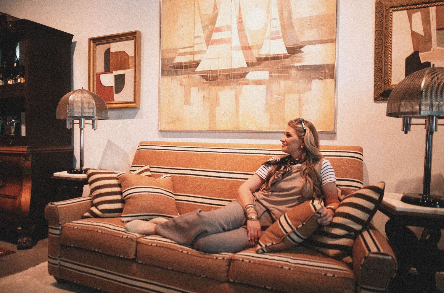 Woman relaxing on a striped sofa with decorative pillows, surrounded by lamps and sailboat artwork in a cozy living room.