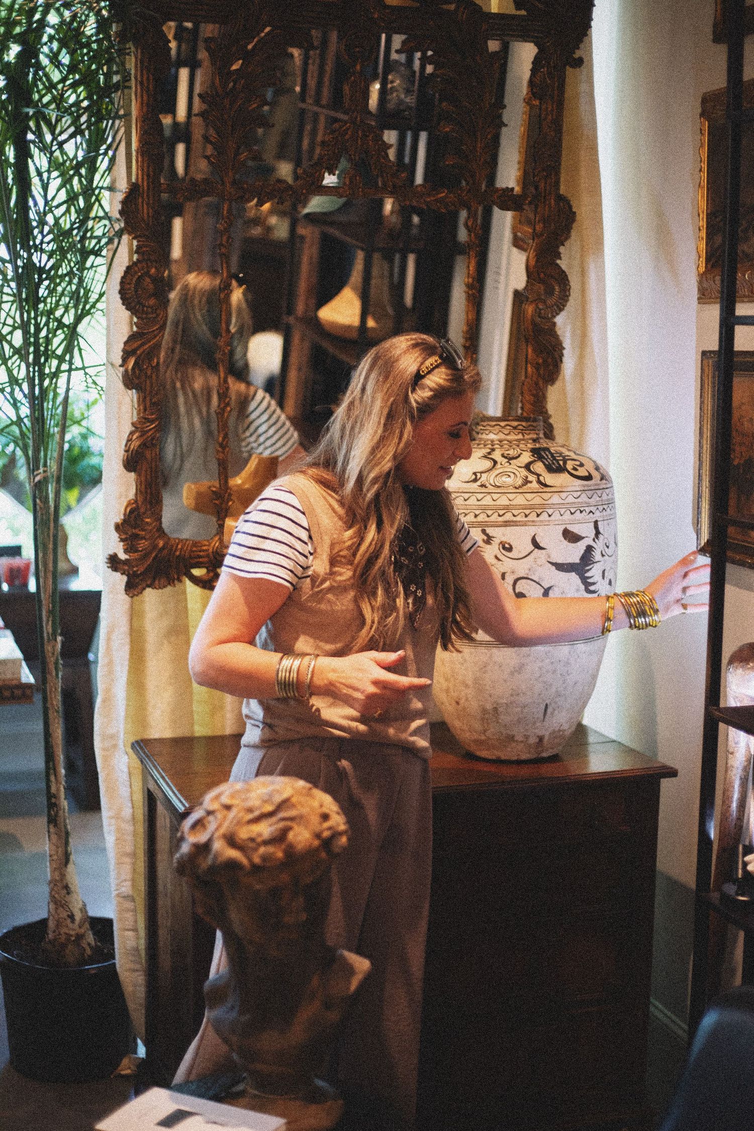 Woman arranging decorative ceramic vase on wooden dresser in eclectic antique shop with ornate gold mirror.