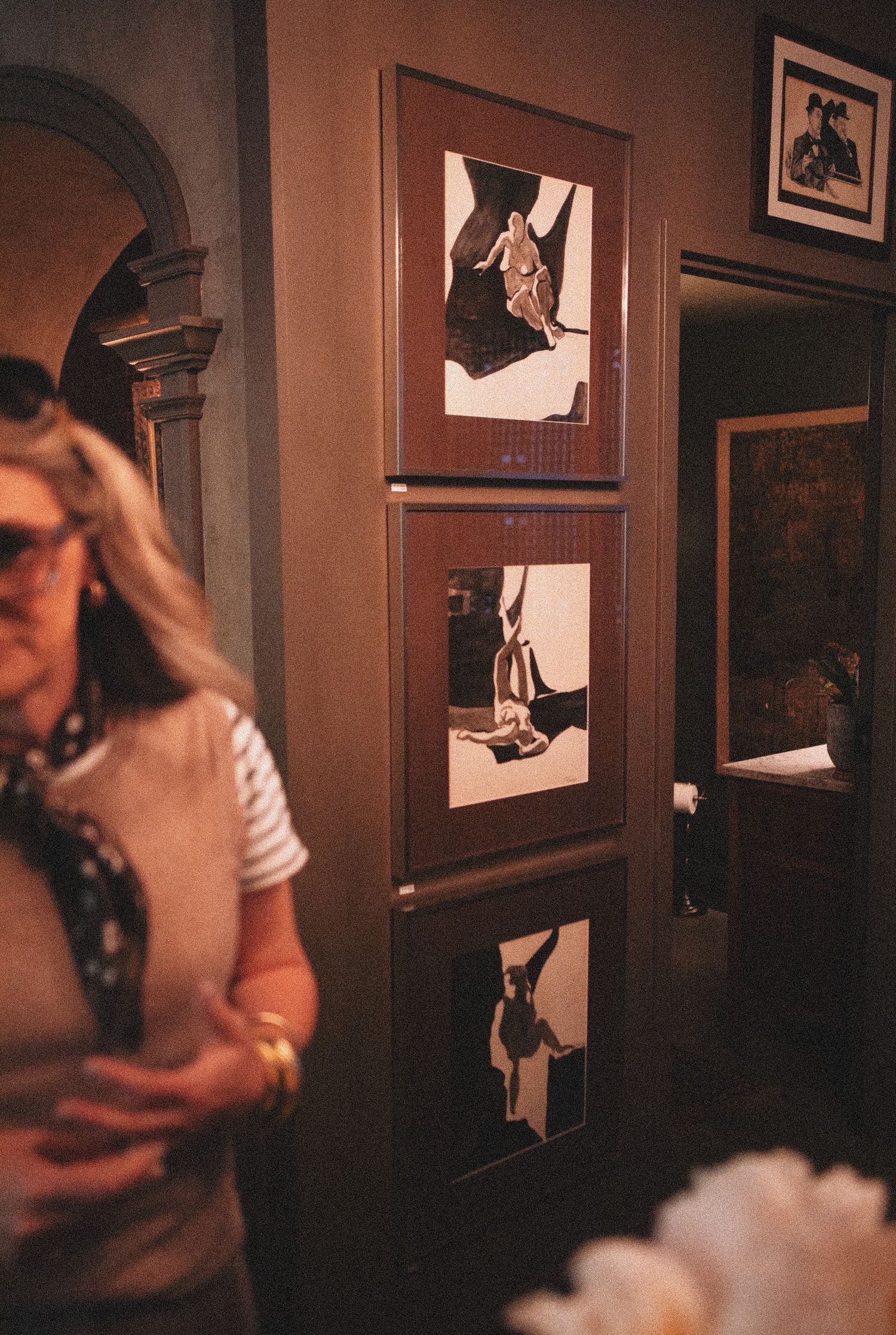 Woman with sunglasses stands near wall displaying three framed black and white photographs in a warmly lit interior.