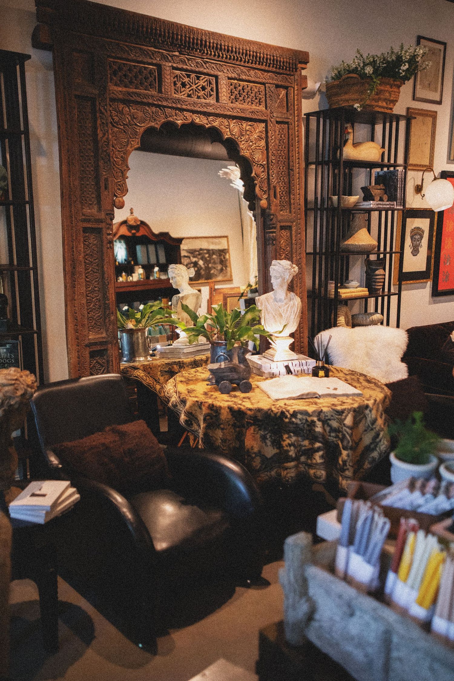 Eclectic living room with ornate carved wood mirror, marble bust, tapestry table, leather chair, and decorative shelving.
