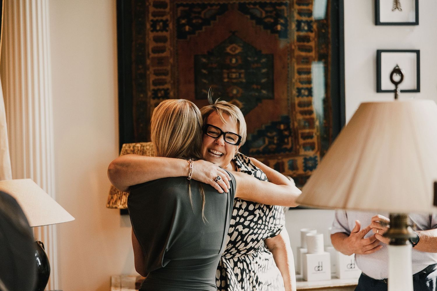Two women sharing a warm hug indoors, smiling joyfully, with a decorative tapestry and lamp in the background.