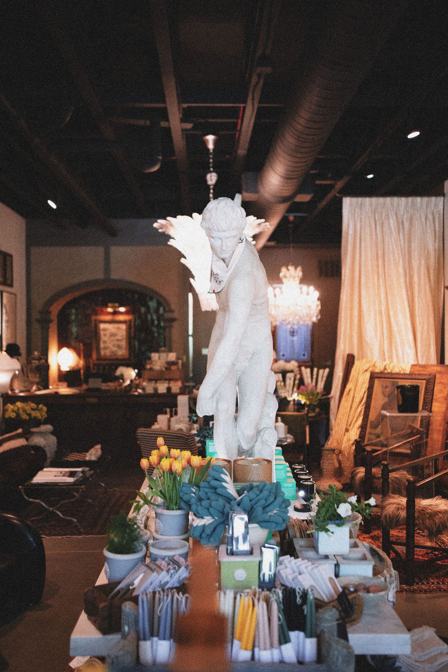 White angel statue centerpiece surrounded by candles, tulips, and decor in an elegant dimly lit boutique interior.
