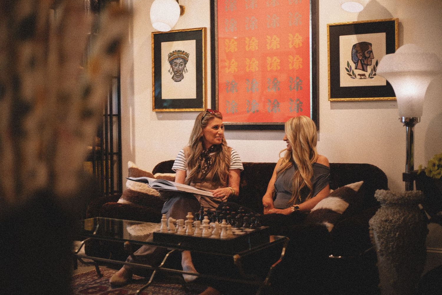 Two women sitting on a dark sofa, reviewing a book in a warmly lit room with eclectic art and a chess set.
