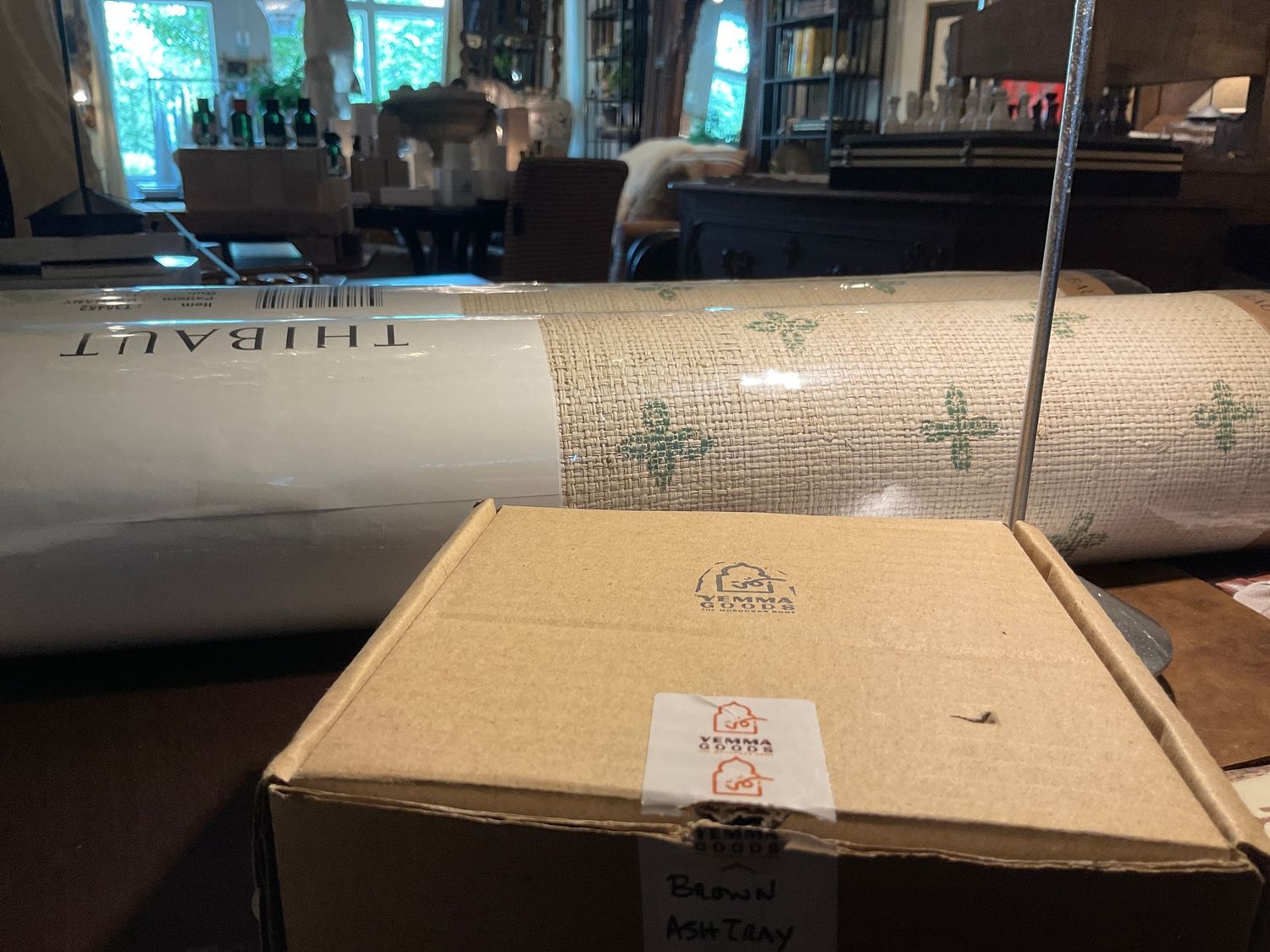 Rolled Thibaut wallpaper and boxed Schumacher fabric samples displayed in a well-lit room with bookshelves.