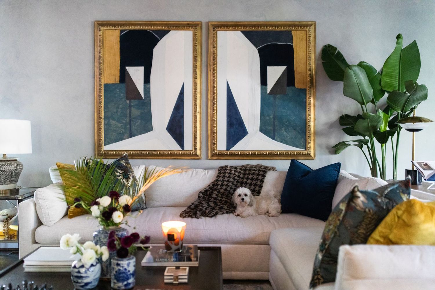 Elegant living room with gold-framed abstract art, white sectional sofa, navy pillows, and a small white dog among lush greenery.