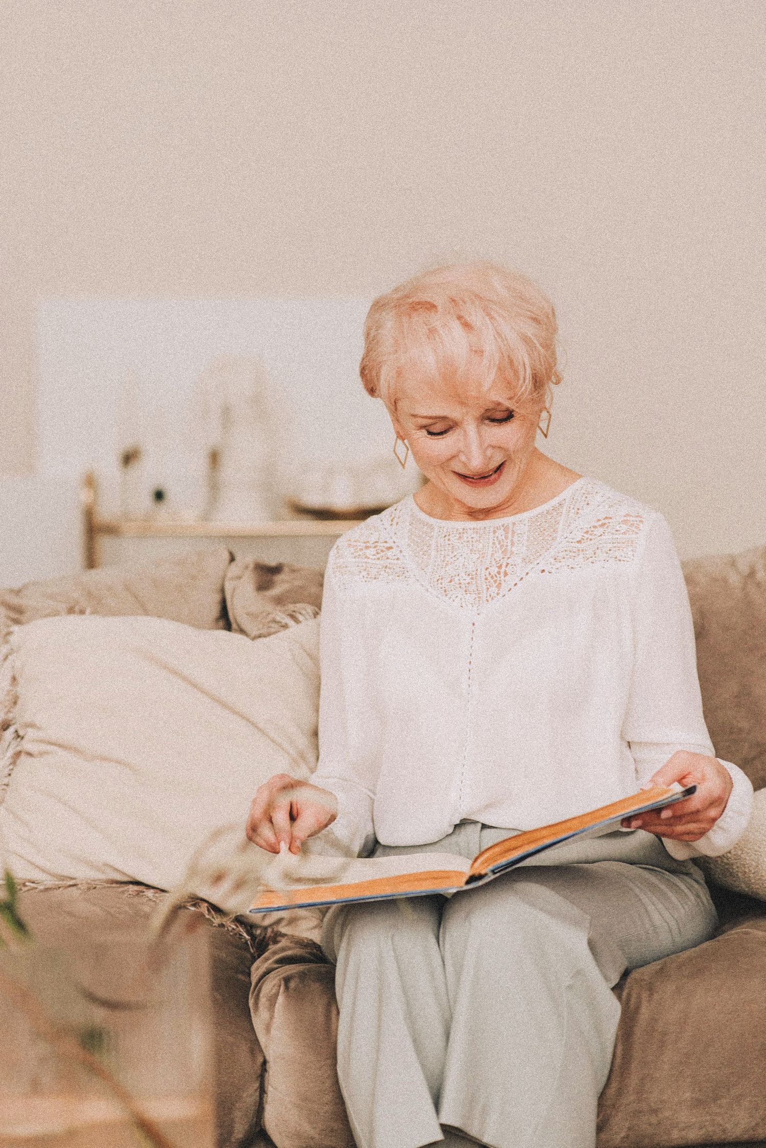Smiling senior woman with short blonde hair reading a book on a cozy sofa in a bright living room.