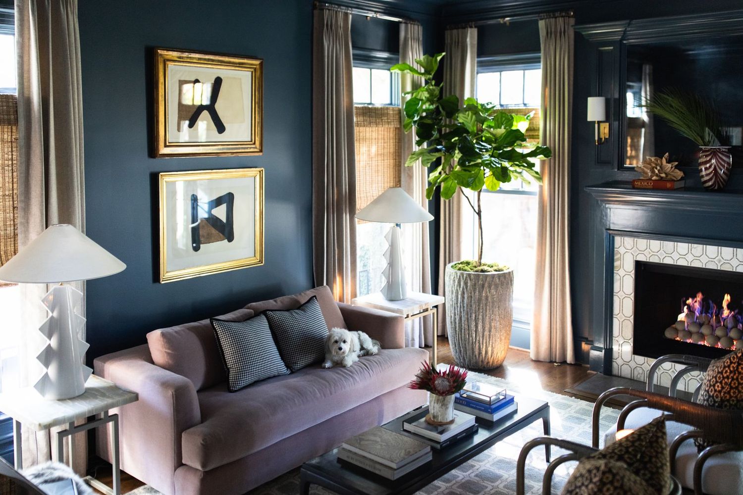 Elegant dark blue living room with pink sofa, gold-framed art, fireplace, and lush fiddle leaf fig tree.