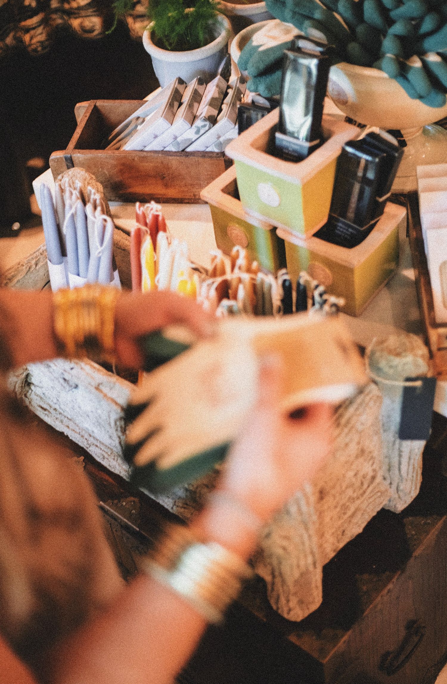 Hands with gold bracelets browse colorful candles and artisan goods displayed in wooden boxes at a bohemian market stall.