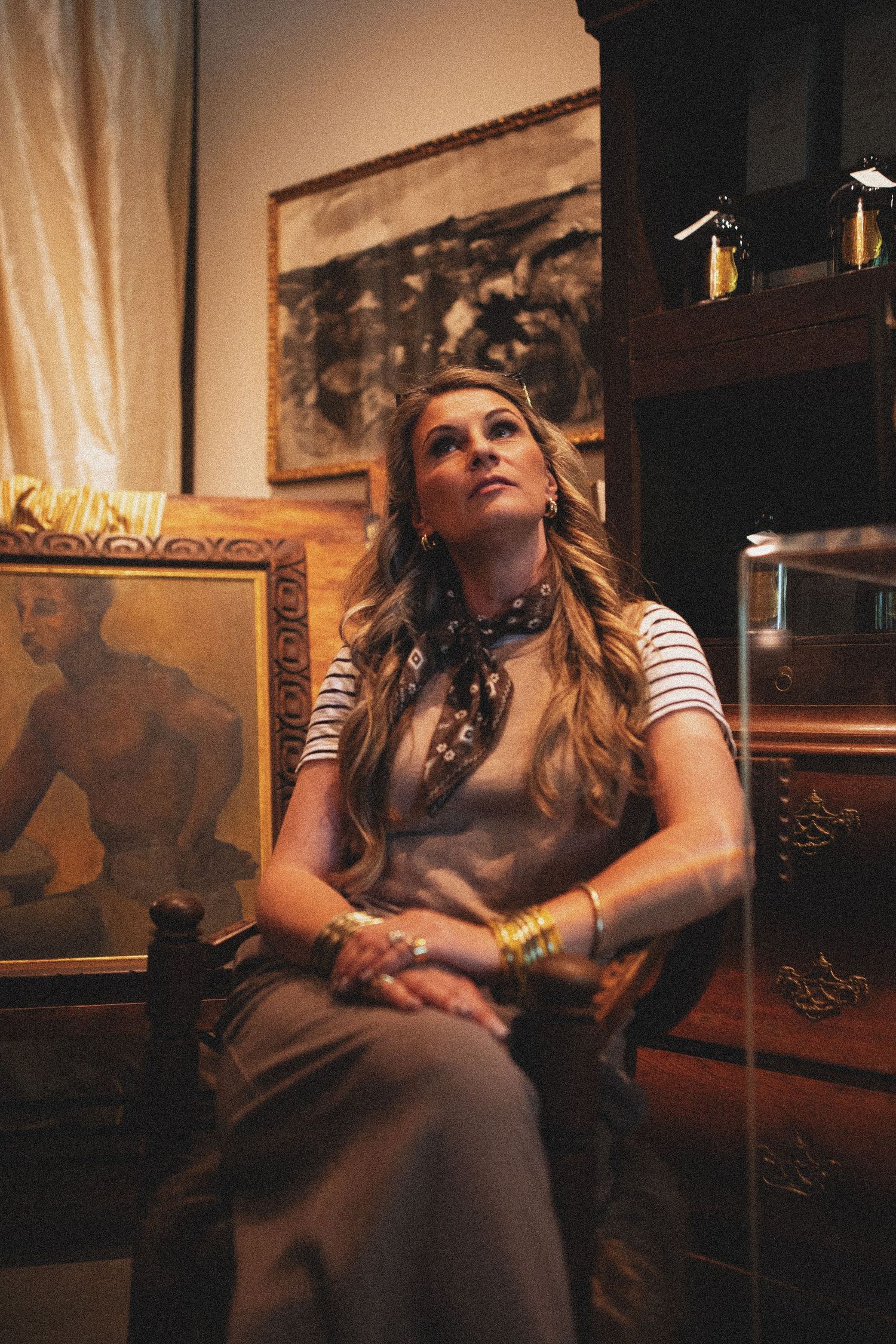 Blonde woman sitting in ornate chair surrounded by antique paintings and wooden furniture in warm-lit room.