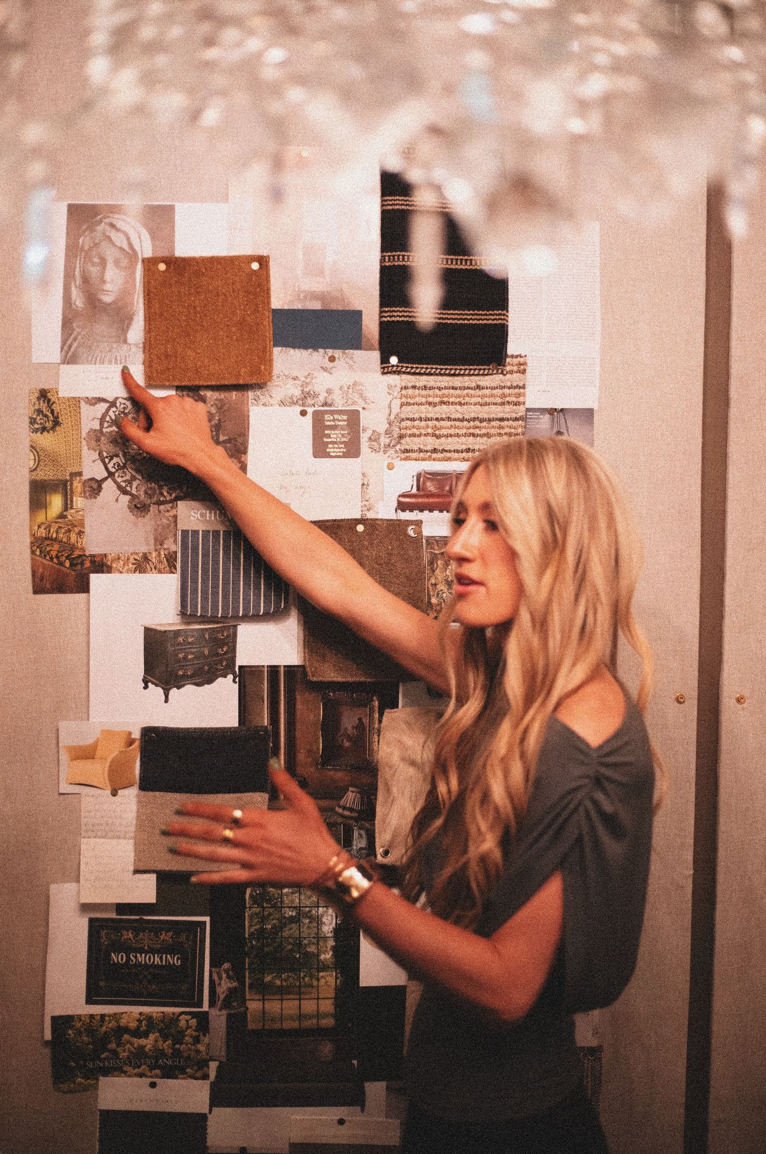 Blonde woman arranging inspiration mood board with fabric swatches and photos on wall.
