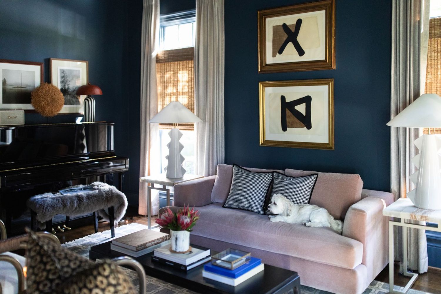 Navy blue living room with pink velvet sofa, gold-framed abstract art, black piano, and a white dog resting on cushions.