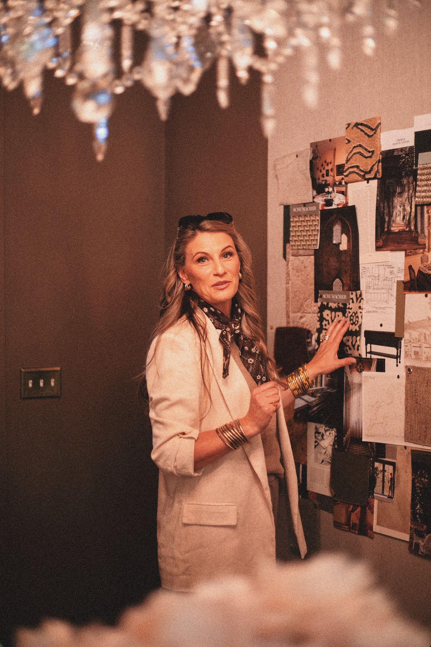 Elegant woman in white blazer and silk scarf gestures at a mood board under a crystal chandelier.