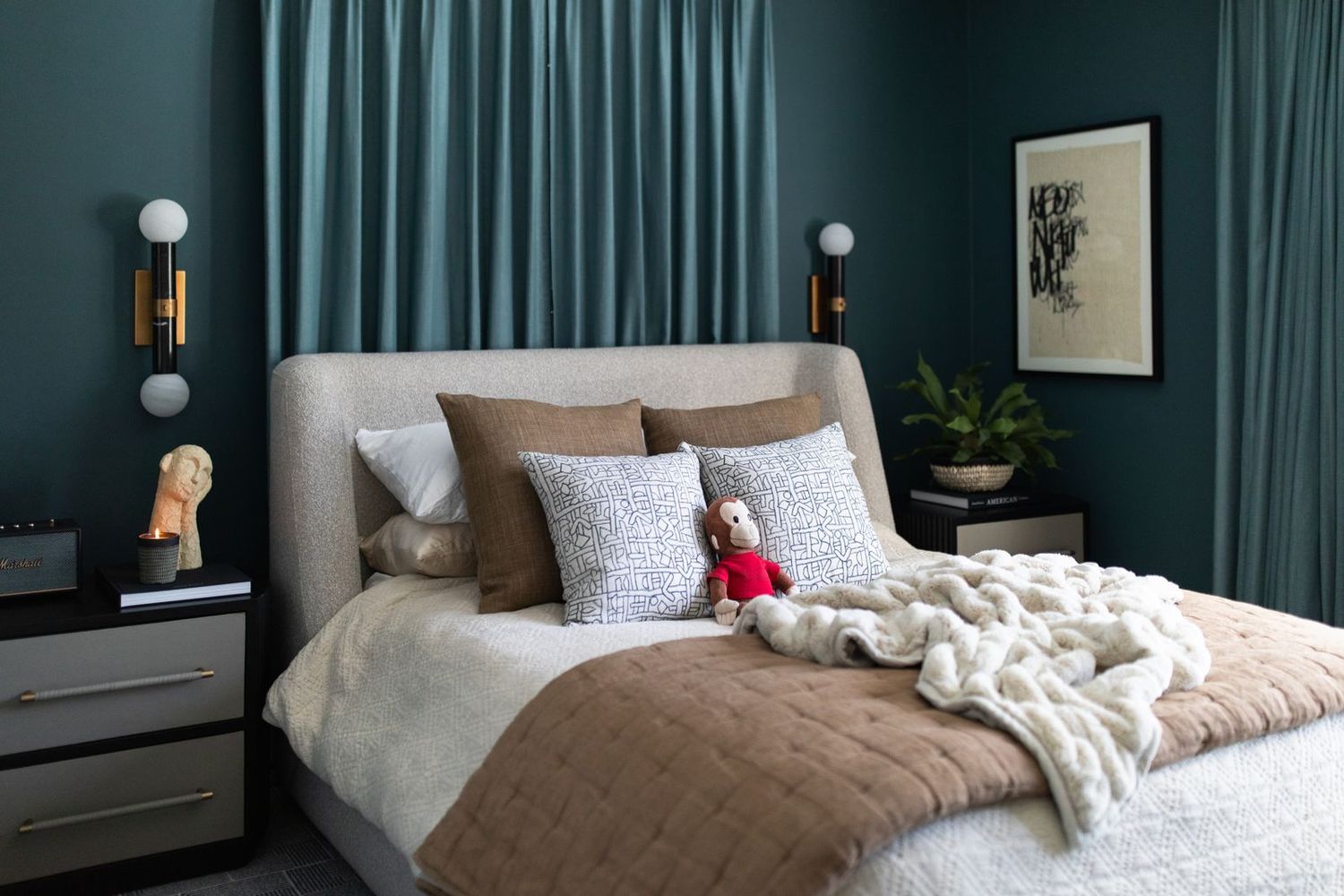 Elegant bedroom with teal walls, upholstered headboard, layered bedding, chunky knit throw, and gold wall sconces.