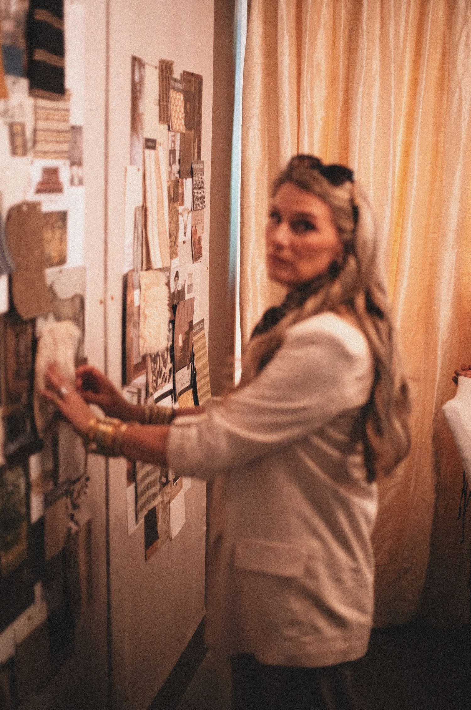 Blonde woman in white blazer pinning items to a cork mood board wall in a warmly lit room.