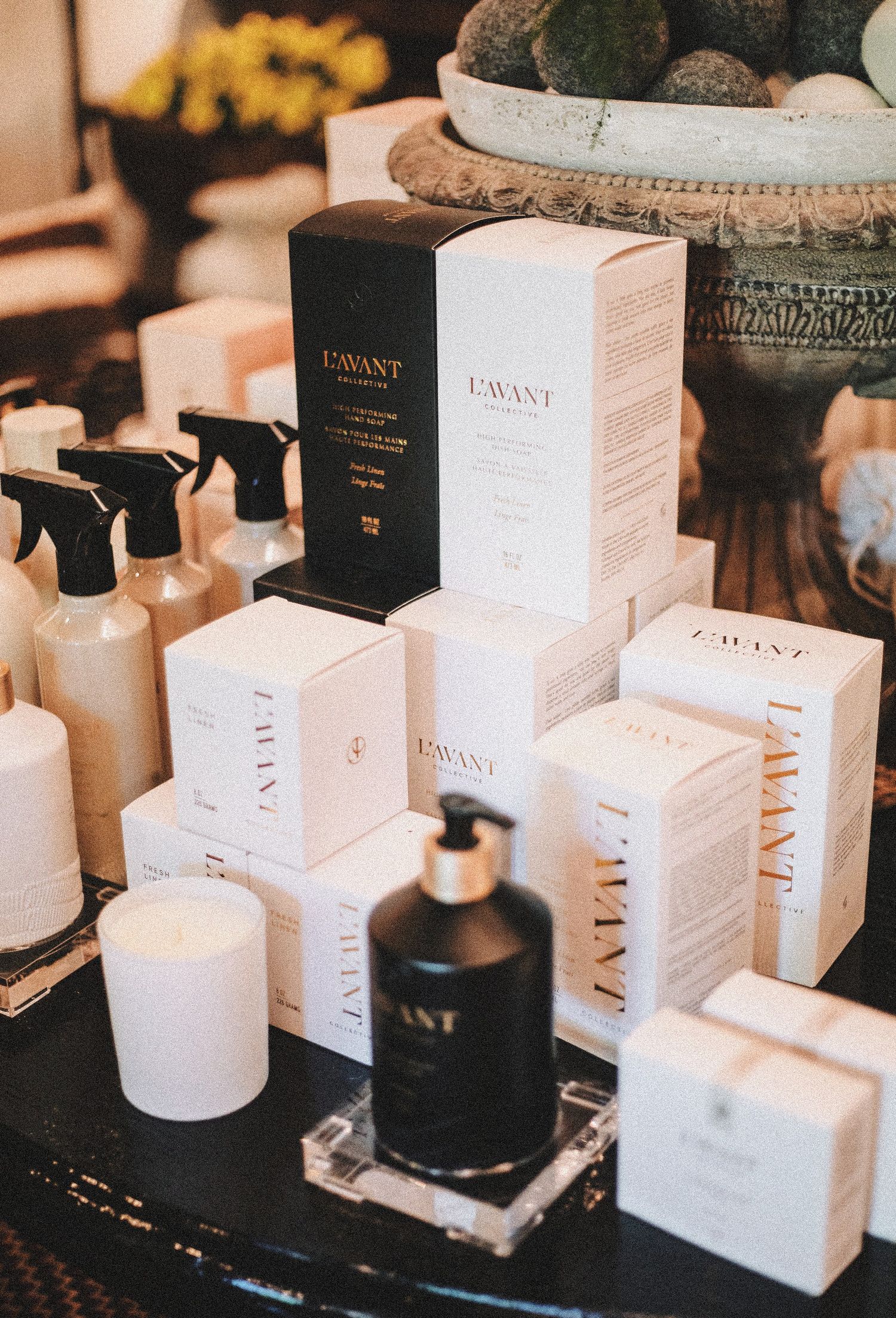 L'Avant Collective cleaning products displayed on a dark table, featuring white boxes, spray bottles, and a black soap dispenser.