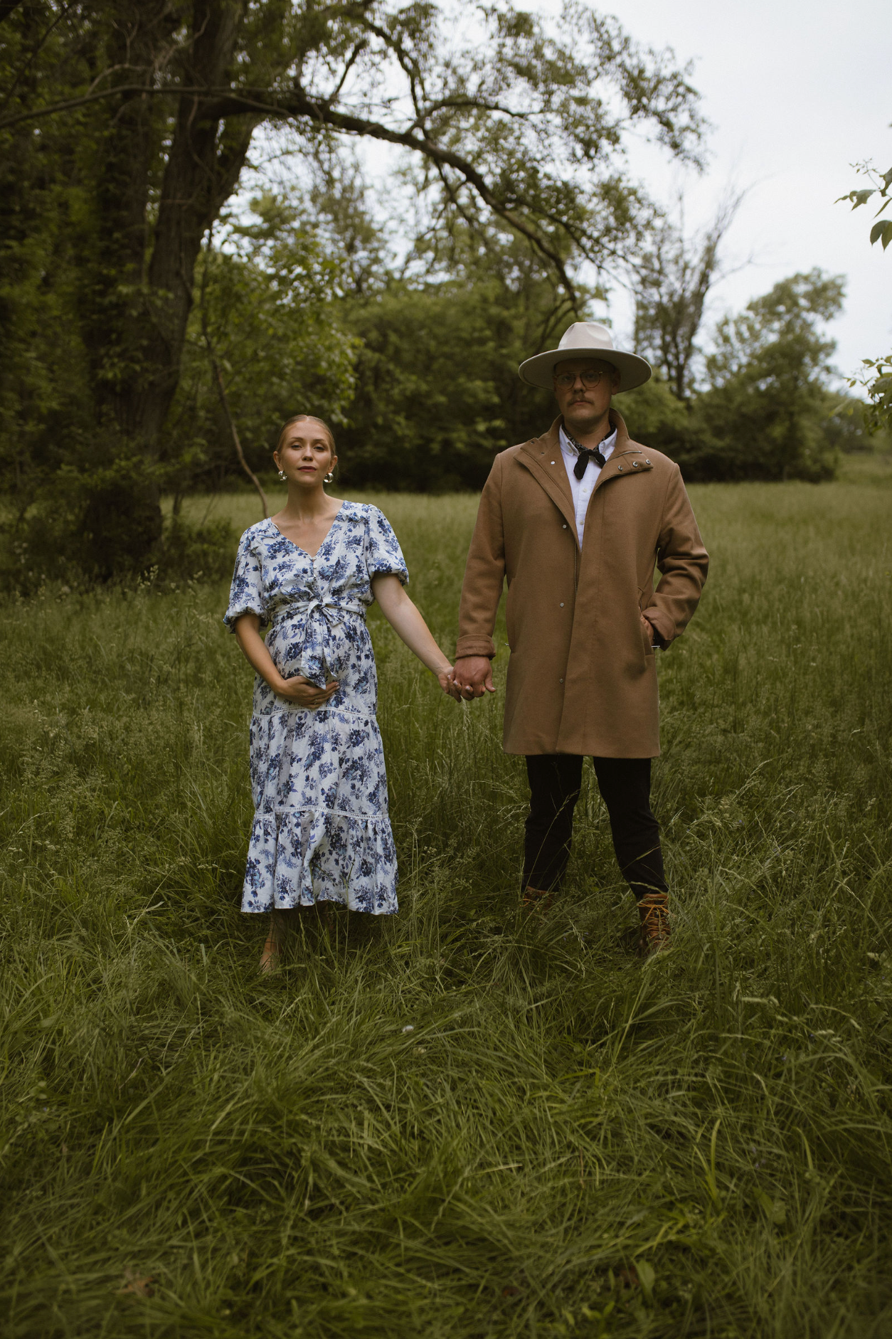 Couple standing in tall grass field wearing vintage-style clothing on an overcast day.